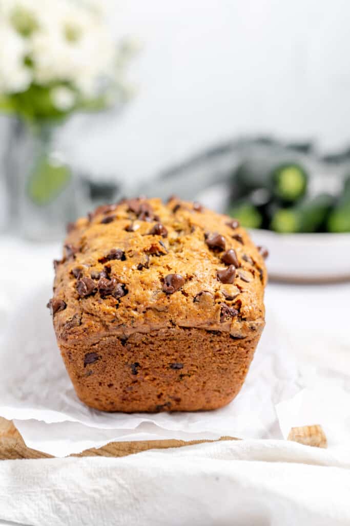 Loaf of chocolate chip zucchini bread on a plate with zucchini piled in the background. 