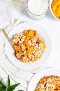 Peach crumble on a plate with a spoon and a cup of milk next to it.