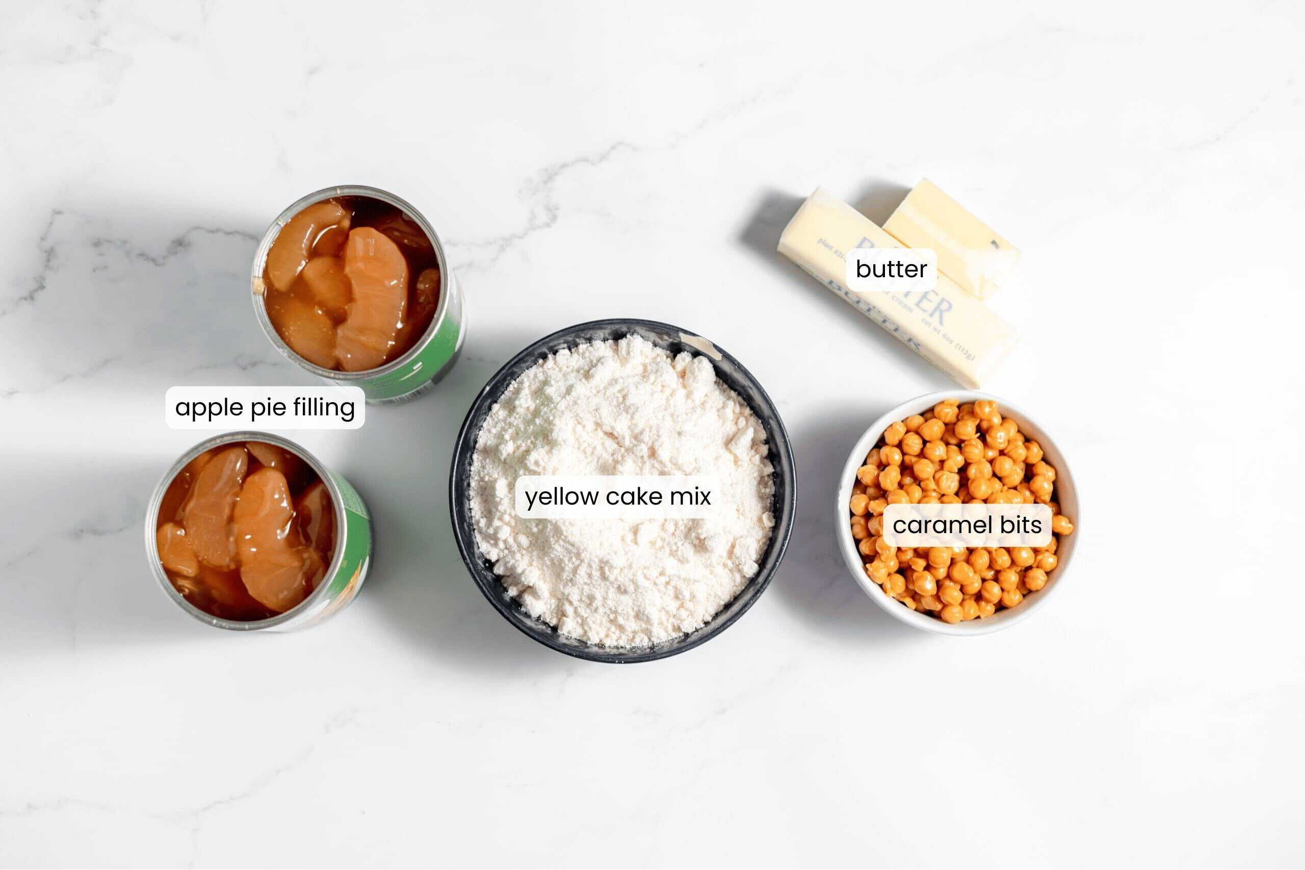 Four ingredients for caramel apple dump cake on a white surface: two cans of apple pie filling, a bowl of yellow cake mix, caramel bits, and two sticks of butter. Labels clearly identify each delicious component.