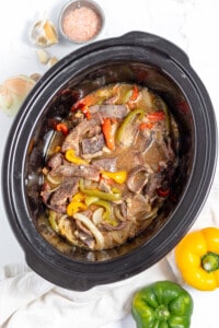 A crock pot full of beef, peppers and onions.