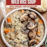 A bowl of creamy mushroom wild rice soup topped with shredded cheese and sliced mushrooms, with a spoon on the side and a small dish of extra cheese nearby for your perfect mushroom wild rice soup experience.