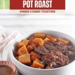 A bowl of slow cooker pot roast with tender chunks of carrots and potatoes sits on a table next to a small dish of savory sauce.