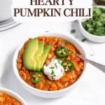 A bowl of turkey pumpkin chili topped with avocado slices, jalapeño, sour cream, and herbs, with a spoon and chopped greens nearby.