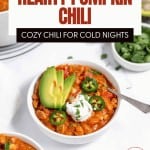 A bowl of turkey pumpkin chili topped with sliced avocado, jalapeños, and sour cream, surrounded by small bowls of chili and chopped herbs. Text overlays promote this hearty turkey pumpkin chili recipe.