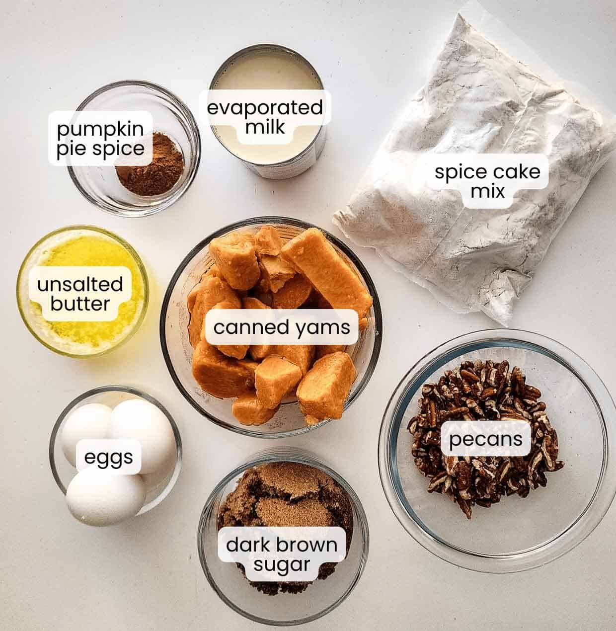 Top-down view of labeled ingredients for a Sweet Potato Dump Cake, including pumpkin pie spice, evaporated milk, spice cake mix, unsalted butter, canned yams, eggs, dark brown sugar, and pecans.