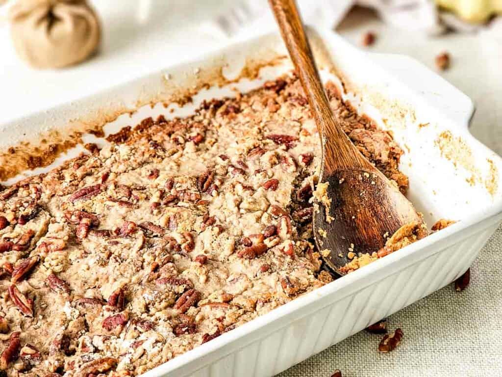 A casserole dish with a wooden spoon sweet potato cake with pecans.