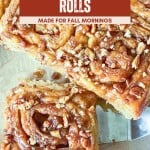 Close-up of apple cinnamon rolls topped with chopped nuts on a tray, with a sign reading "Apple Cinnamon Rolls Made for Fall Mornings." These warm, gooey apple cinnamon rolls are perfect for cozy autumn breakfasts.