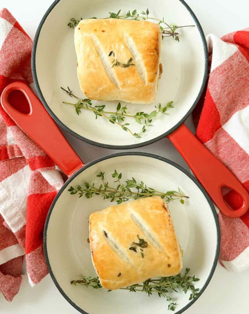 Two pans with two sausage roll pastries in them with sprigs of thyme.
