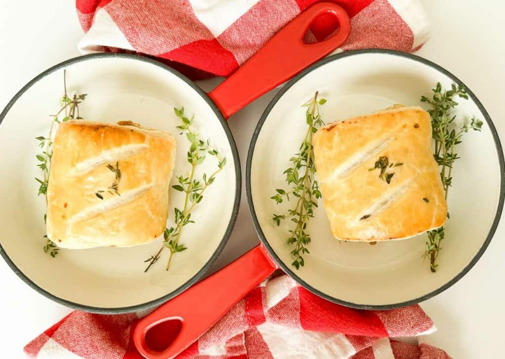 Two sausage rolls with thyme on a red and white checkered tablecloth.