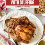 Oven roasted turkey stuffed with classic stuffing sits on a white platter, surrounded by savory gravy, warm bread rolls, and a casserole dish on a white table.