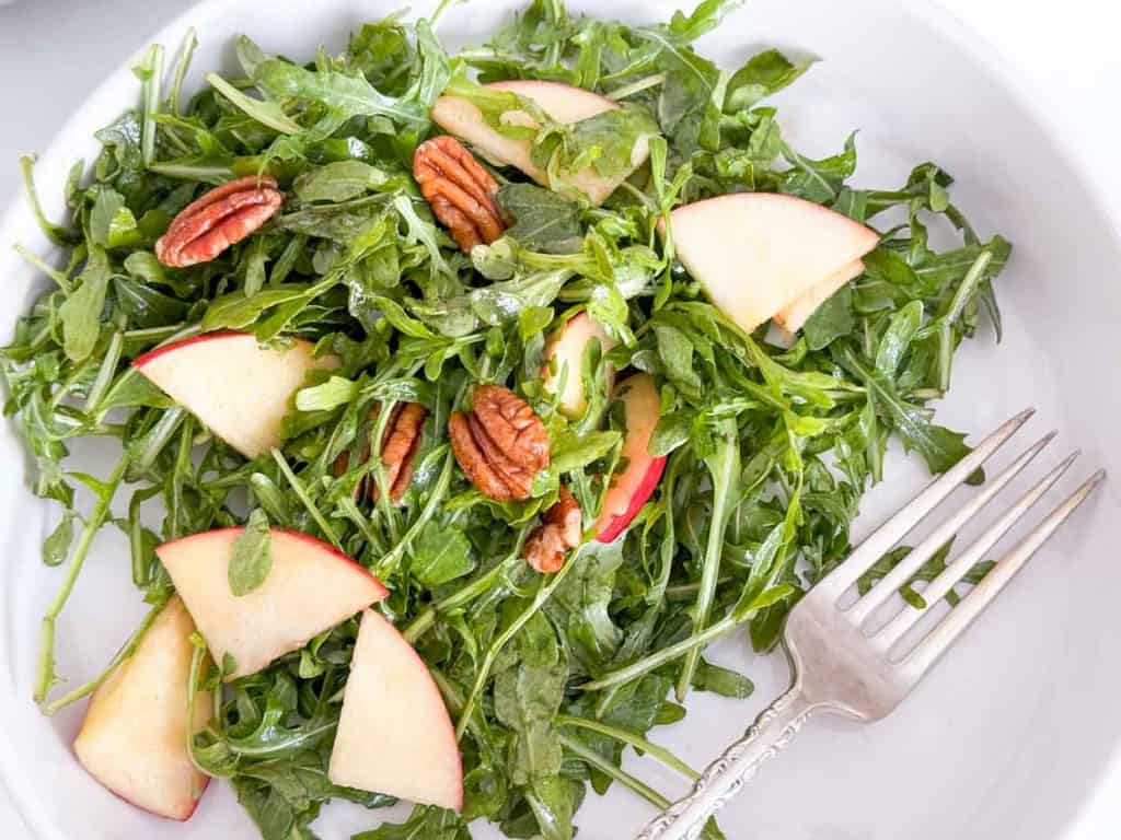 A plate of arugula salad with apples and pecans. The arugula salad is enhanced with the fresh flavors of apples and the delightful crunch of pecans.