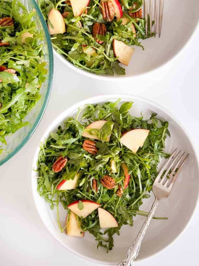 Two bowls of arugula salad with apples and pecans.