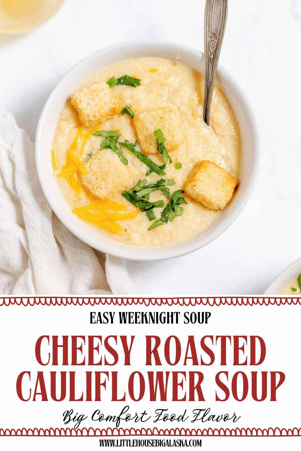 Bowl of creamy roasted cauliflower soup topped with croutons, shredded cheese, and herbs, with a spoon in the bowl. Text below reads “Cheesy Roasted Cauliflower Soup.”.
