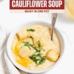 A bowl of cheesy Roasted Cauliflower Soup topped with croutons, shredded cheese, and chopped herbs, with a spoon inside the bowl.