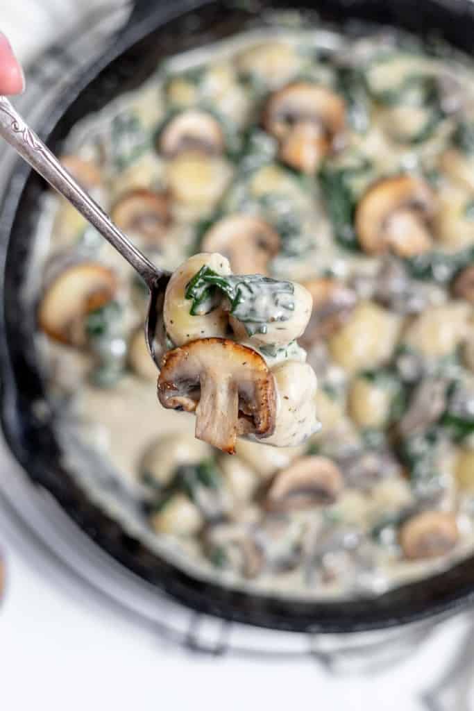 A person is holding a spoon full of spinach and mushrooms in a skillet.