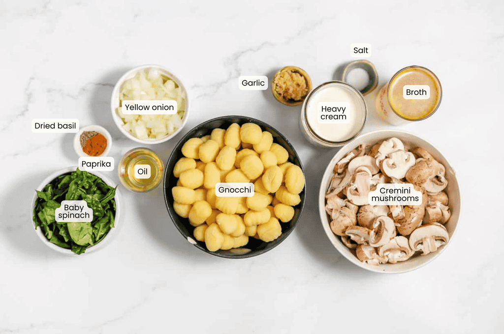 Ingredients on a white surface: gnocchi, cremini mushrooms, baby spinach, yellow onion, garlic, heavy cream, broth, oil, paprika, dried basil, and salt in labeled bowls and containers&mdash;everything you need to make a delicious Mushroom Gnocchi dish.