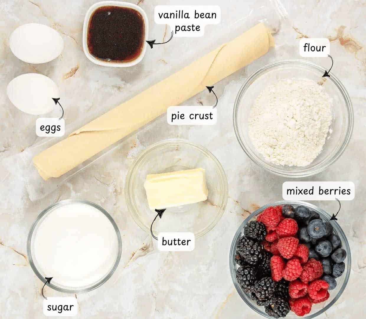 Ingredients for a custard pie are arranged on a surface: pie crust, eggs, vanilla bean paste, flour, sugar, butter, and a bowl of mixed berries including blackberries, raspberries, and blueberries.