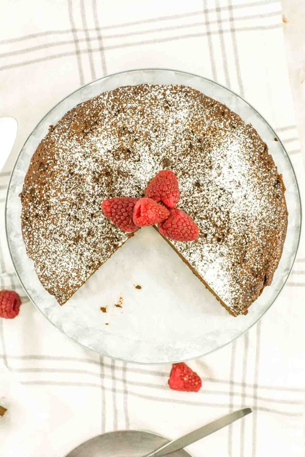 A single slice of flourless chocolate cake topped with raspberries and powdered sugar, with more cake and a slice in the background.