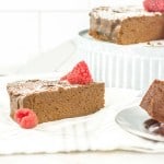 A single slice of flourless chocolate cake topped with raspberries and powdered sugar, with more cake and a slice in the background.