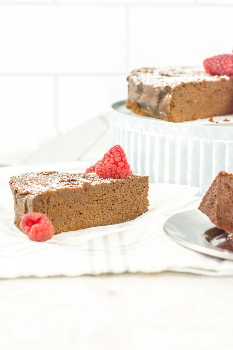 A single slice of flourless chocolate cake topped with raspberries and powdered sugar, with more cake and a slice in the background.