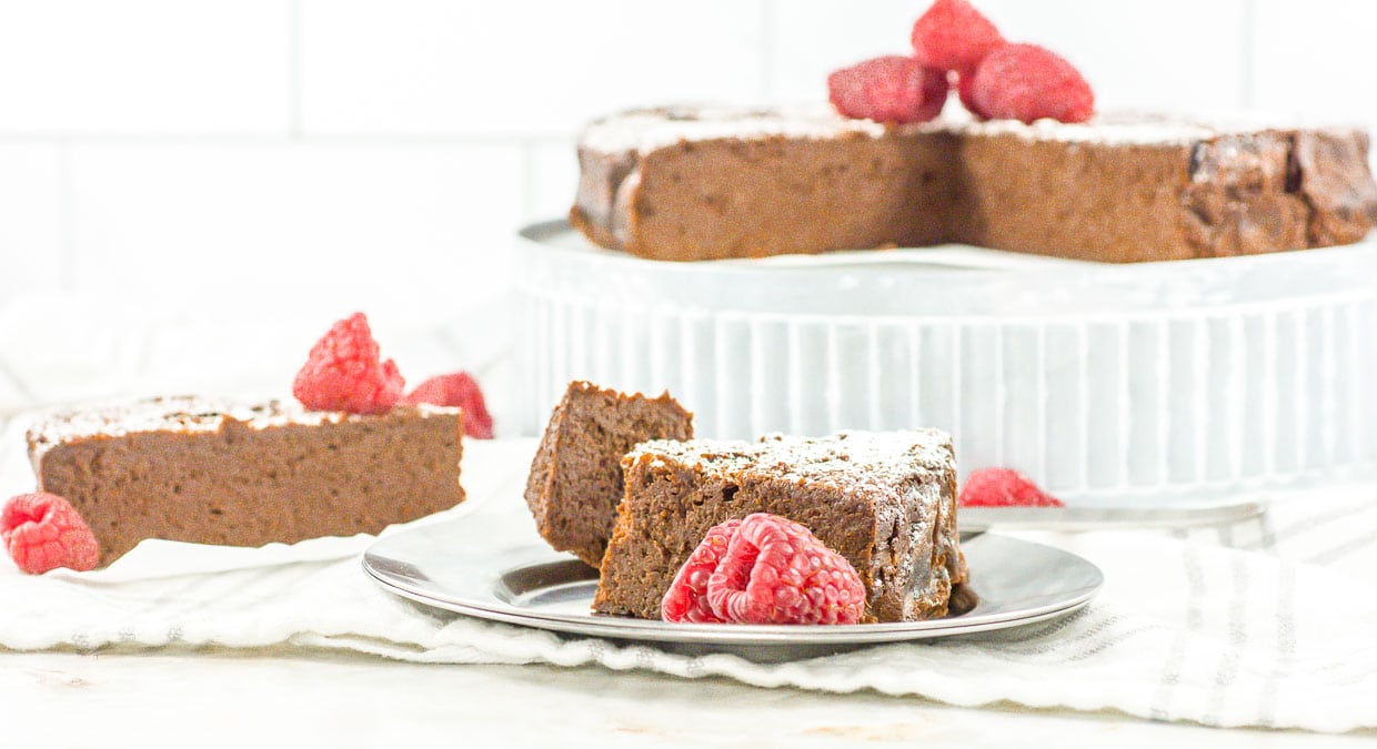 A single slice of flourless chocolate cake topped with raspberries and powdered sugar, with more cake and a slice in the background.