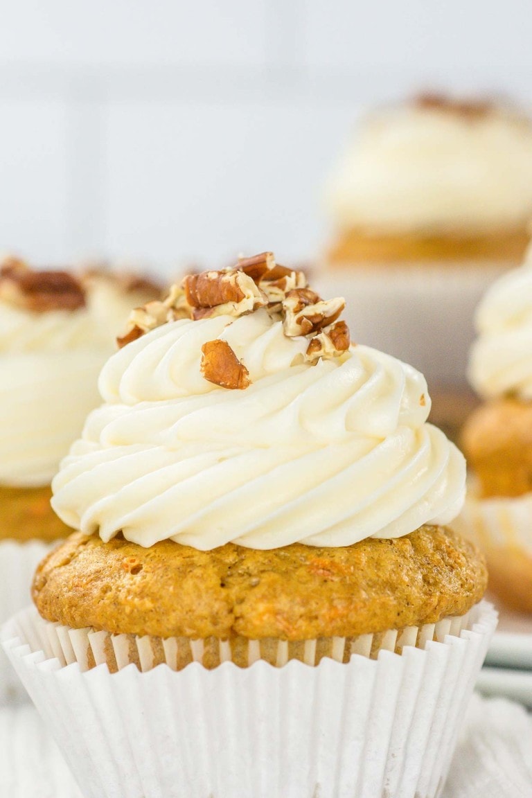 A close-up of a carrot cake cupcake topped with cream cheese frosting and chopped walnuts.