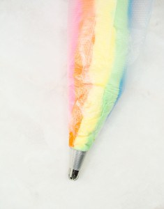 A pastry bag filled with rainbow-colored frosting, with its metal tip visible, against a white background.