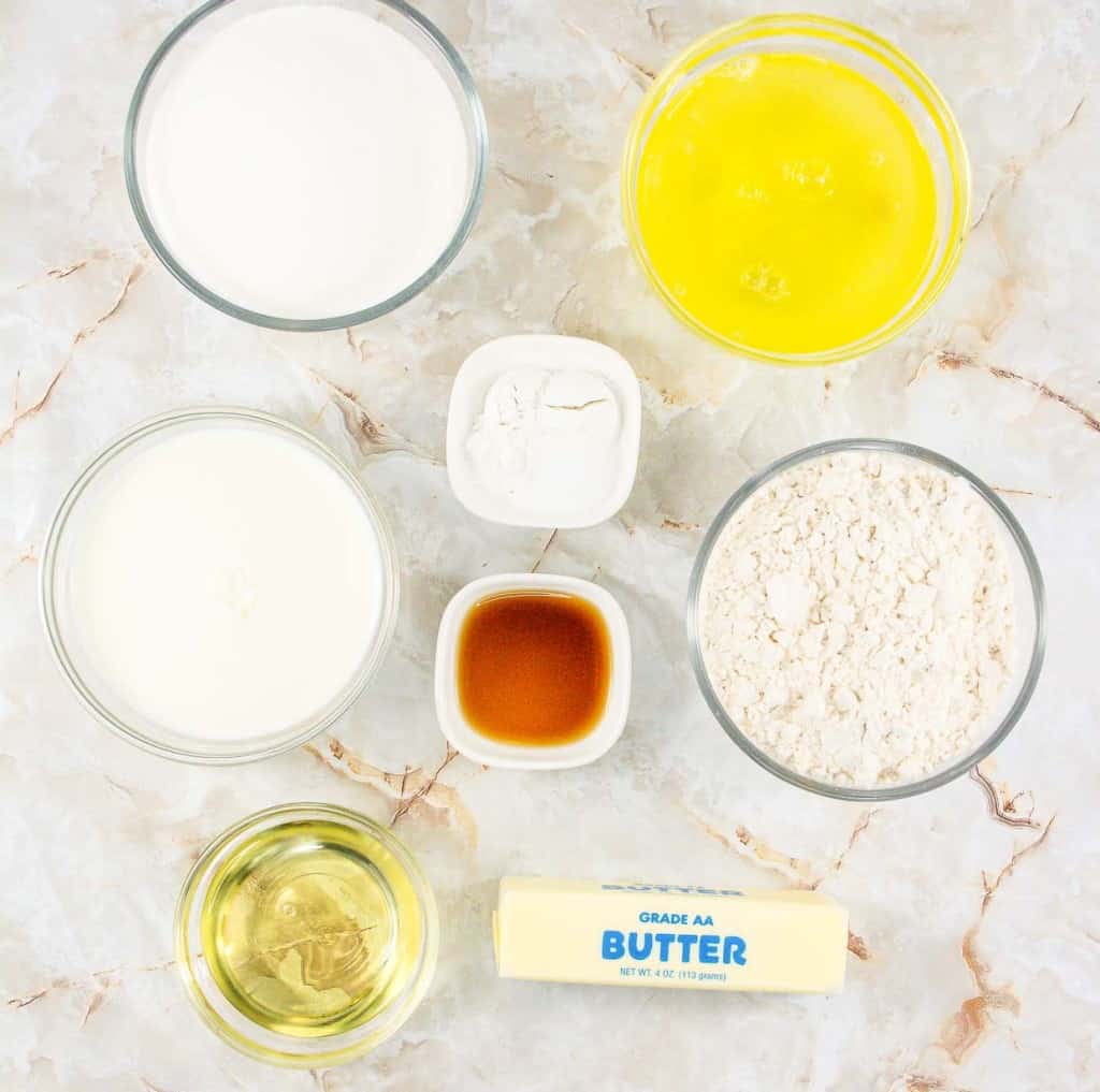 Ingredients for baking arranged on a marble countertop, including flour, melted butter, milk, oil, baking powder, and vanilla extract.