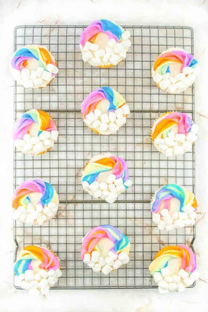 A cooling rack displays several cupcakes each adorned with a colorful rainbow frosting and a cluster of mini marshmallows.