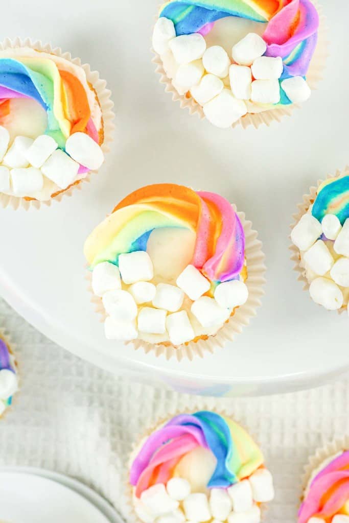 Colorful rainbow frosting adorns cupcakes surrounded by mini marshmallows, presented on a white plate with additional cupcakes in the background.