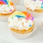 A plate of cupcakes decorated with white frosting, rainbow-colored accents, and mini marshmallows.
