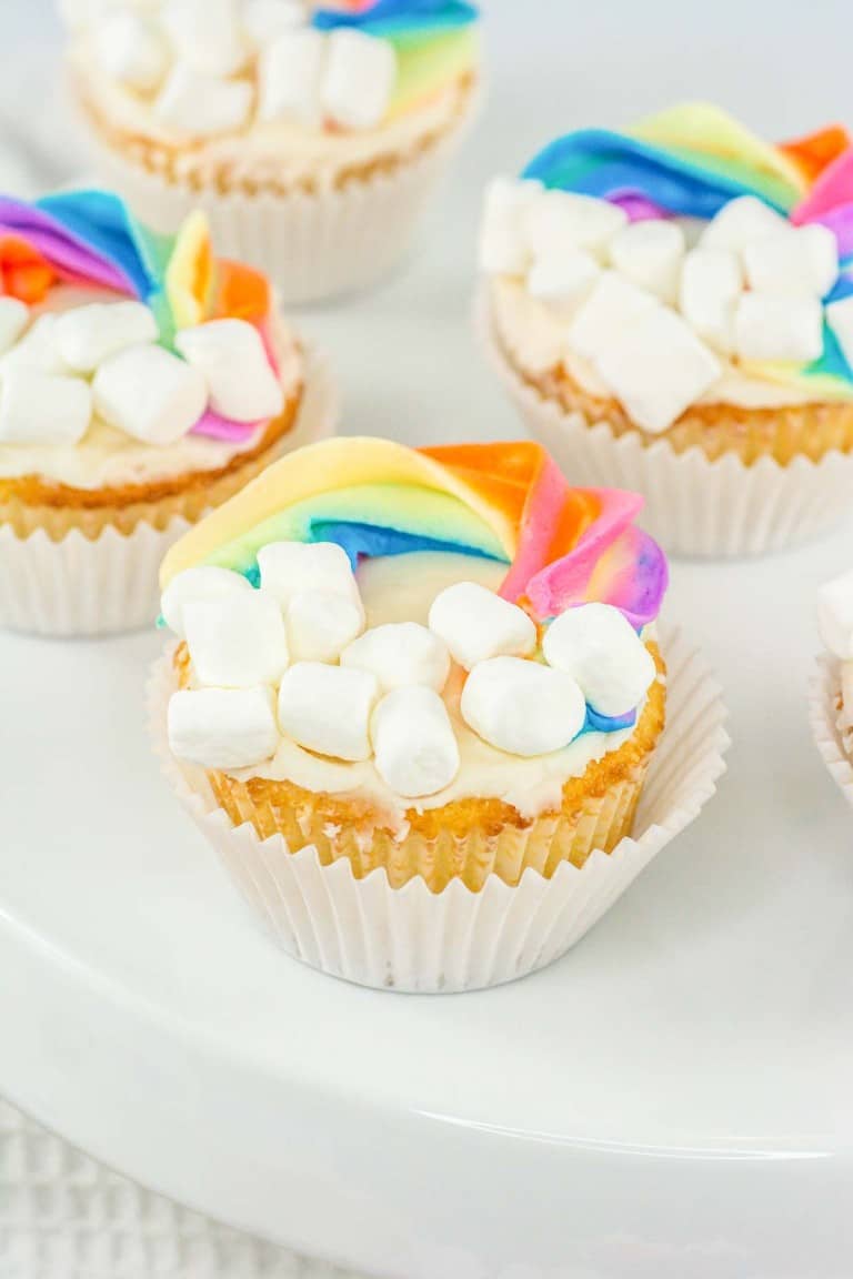 A plate of cupcakes decorated with white frosting, rainbow-colored accents, and mini marshmallows.