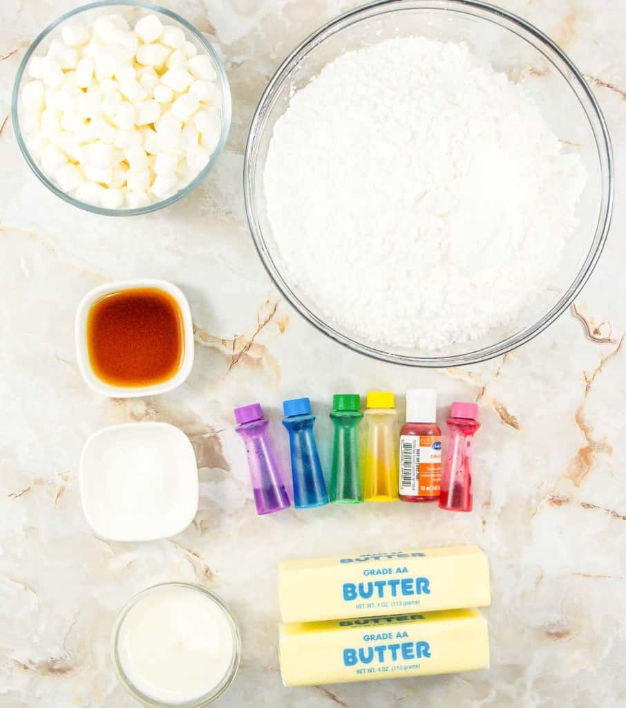 Ingredients for baking laid out on a marble surface, including marshmallows, flour, vanilla extract, food coloring, powdered sugar, and butter.