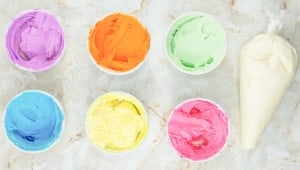 Seven bowls of variously colored frosting (purple, orange, green, blue, yellow, and pink) aligned next to a piping bag filled with white frosting, all set on a marble surface.