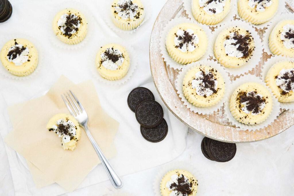 A display of mini cookies and cream cheesecakes with cream frosting and cookie crumble toppings, accompanied by a fork and some whole cookies on a napkin.
