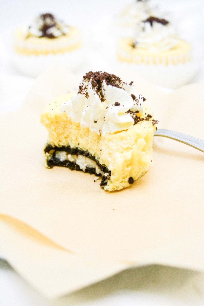 A display of mini cookies and cream cheesecakes with cream frosting and cookie crumble toppings, accompanied by a fork and some whole cookies on a napkin.