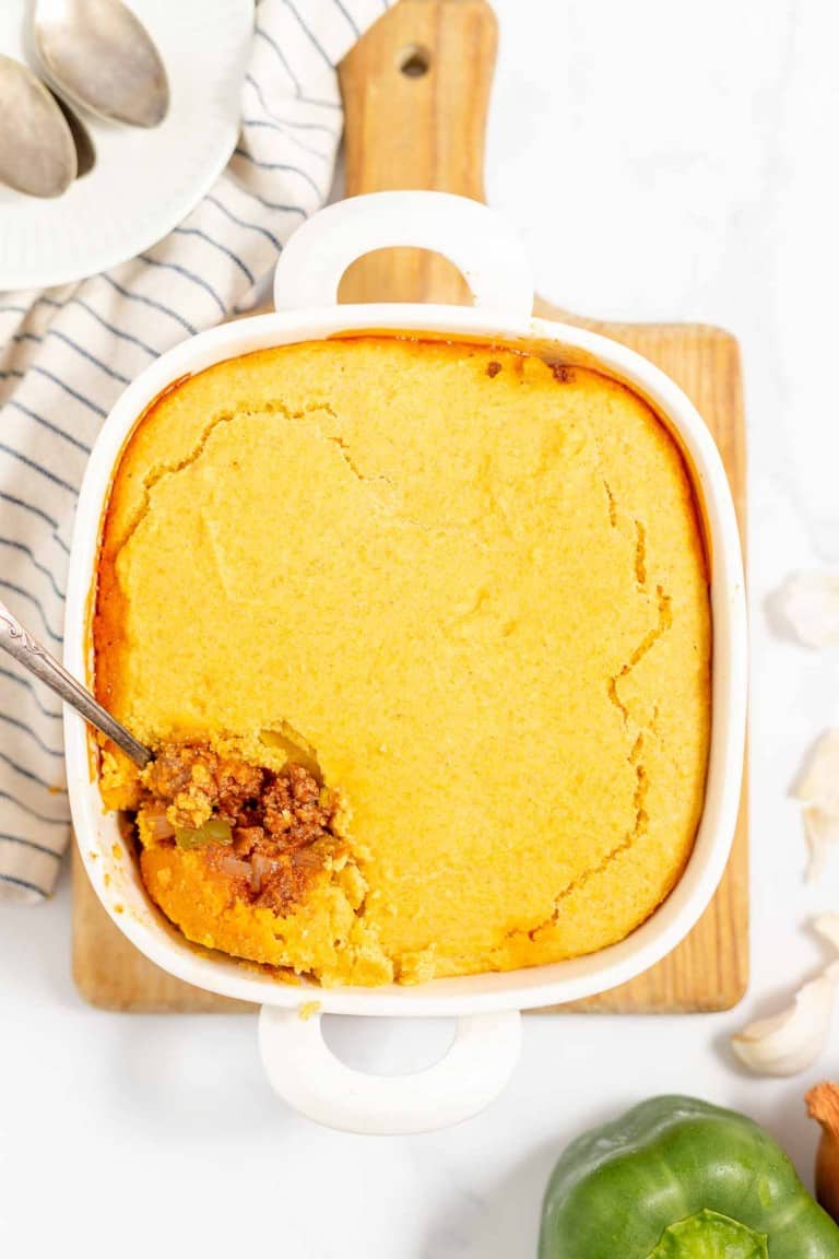 A baking dish with cornbread topping over a layer of chili, partially scooped out, placed on a wooden board with a striped cloth and vegetables nearby.