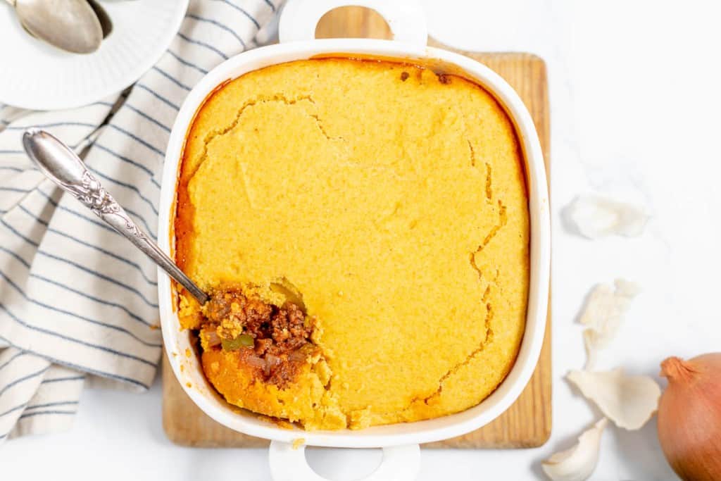 A casserole dish with a golden cornbread crust on top and a spoon scooping out the meat filling.