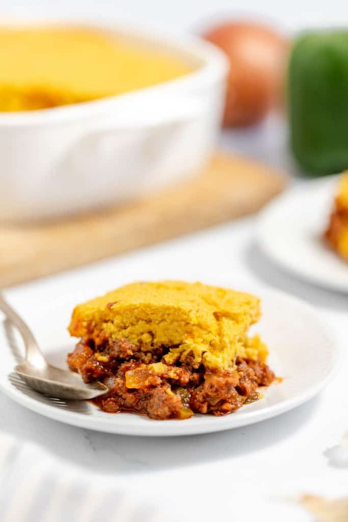 A slice of cornbread-topped chili pie served on a white plate with a fork, with a green bell pepper and ingredients in the background.