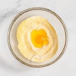 A bowl of flour with a cracked egg in the center on a marble countertop.