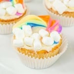 A cupcake decorated with a rainbow icing arch and marshmallows to resemble clouds on a white surface.