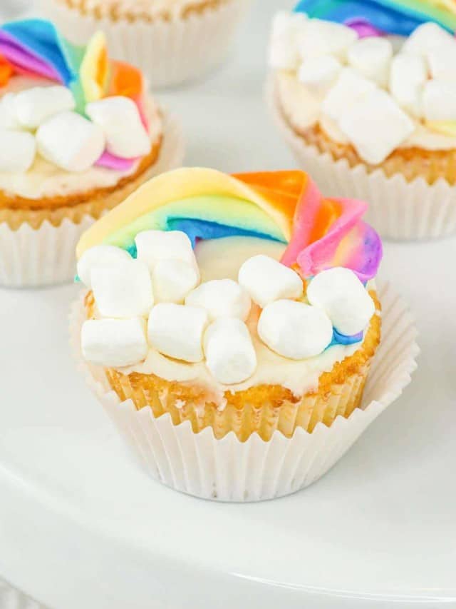 A cupcake decorated with a rainbow icing arch and marshmallows to resemble clouds on a white surface.