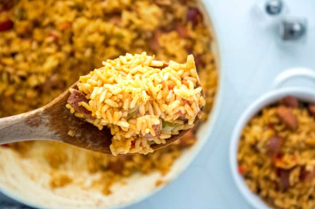 A wooden spoon lifting a serving of vibrant, cooked jollof rice with visible pieces of vegetables, over a pot of the *one-pot cajun* rice dinner in the background.