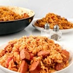 A plate of one-pot jambalaya with sausage pieces in front, with more jambalaya in a pan and salt and pepper shakers in the background.