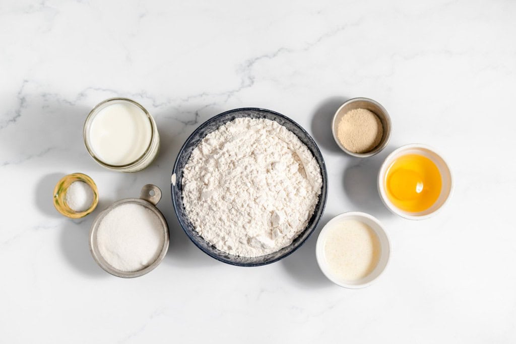 Overhead view of copycat Texas Roadhouse rolls baking ingredients on marble surface, including flour, milk, sugar, oil, egg, and yeast.