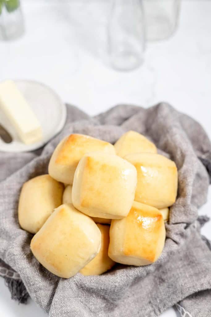 A pile of freshly baked copycat Texas Roadhouse rolls with a glossy top, resting on a gray cloth, with butter and glasses in the background on a white surface.