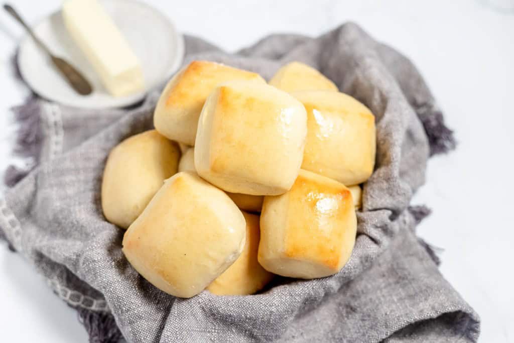 Freshly baked copycat Texas Roadhouse rolls on a cloth napkin with butter in the background, displayed on a white marble surface.