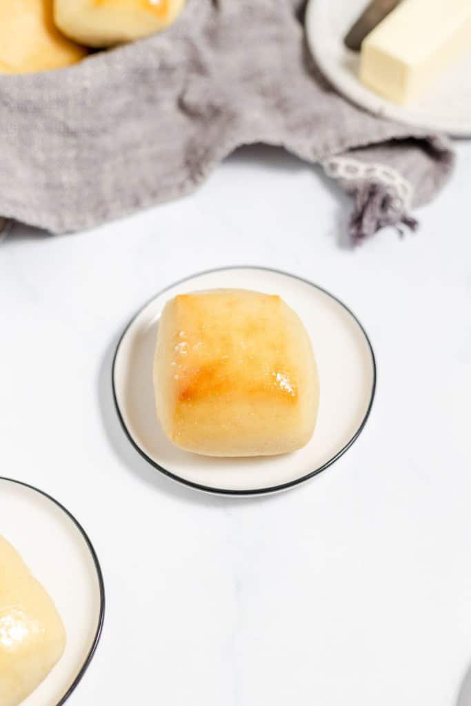 A freshly baked copycat Texas Roadhouse roll glazed with butter on a small white plate, with a stick of butter and a cloth napkin in the background.