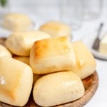 Freshly baked copycat Texas Roadhouse rolls on a wooden plate with a butter dish and glass in the background.