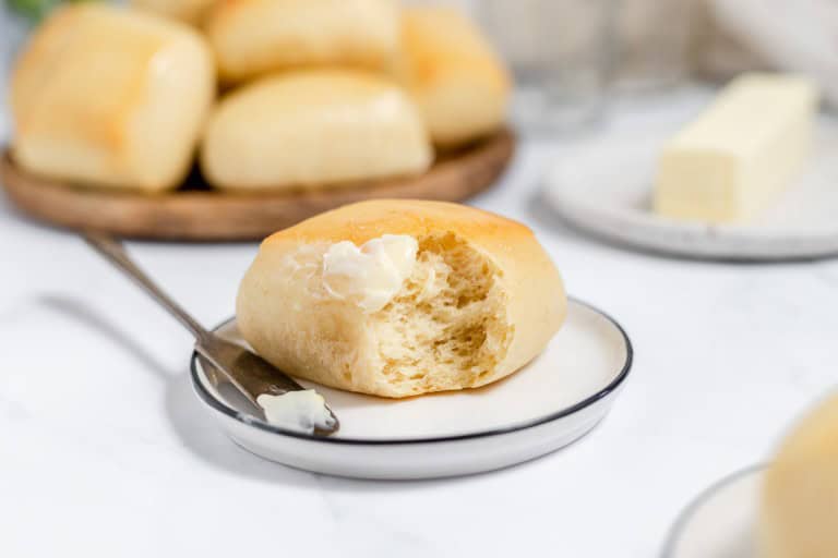 A fresh copycat Texas Roadhouse roll with butter on a small plate, additional rolls and a butter dish in the background on a marble surface.